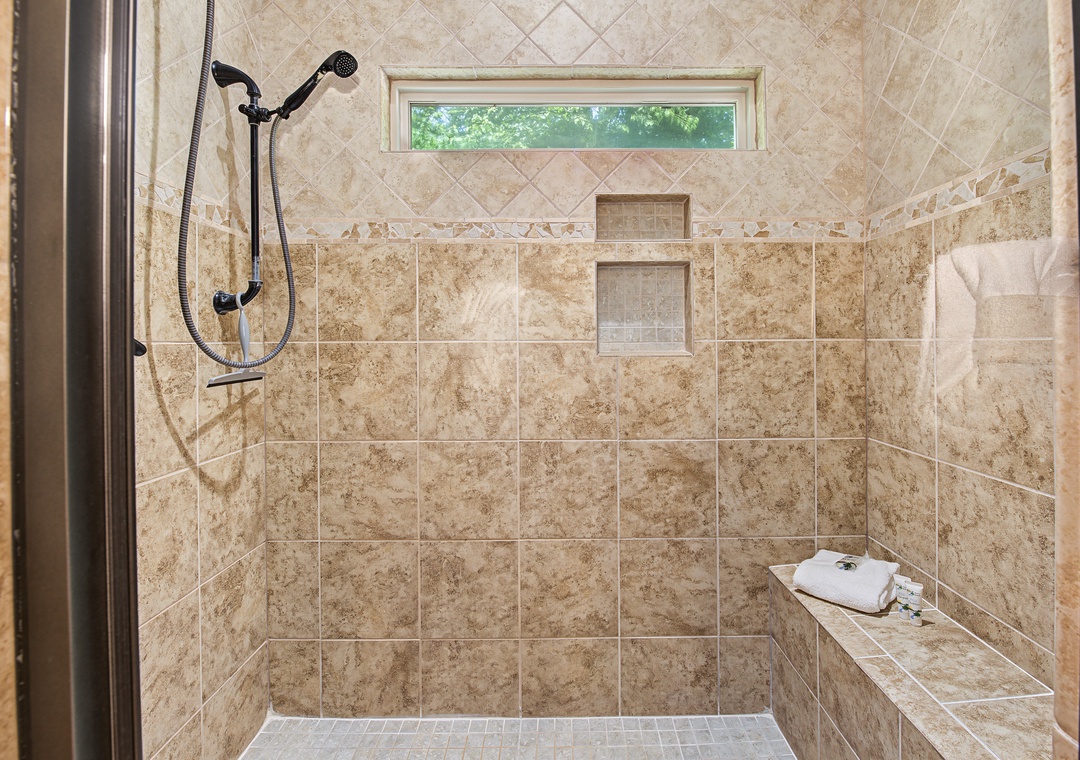 Step into your private spa-like retreat. This spacious walk-in shower features beautiful travertine tile, a built-in bench for relaxation, dual showerheads, and a window bringing in natural light—pure comfort awaits you.