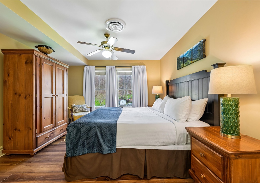 Relax in your peaceful bedroom with plush bedding, warm wooden furnishings, and natural light streaming through garden-view windows.