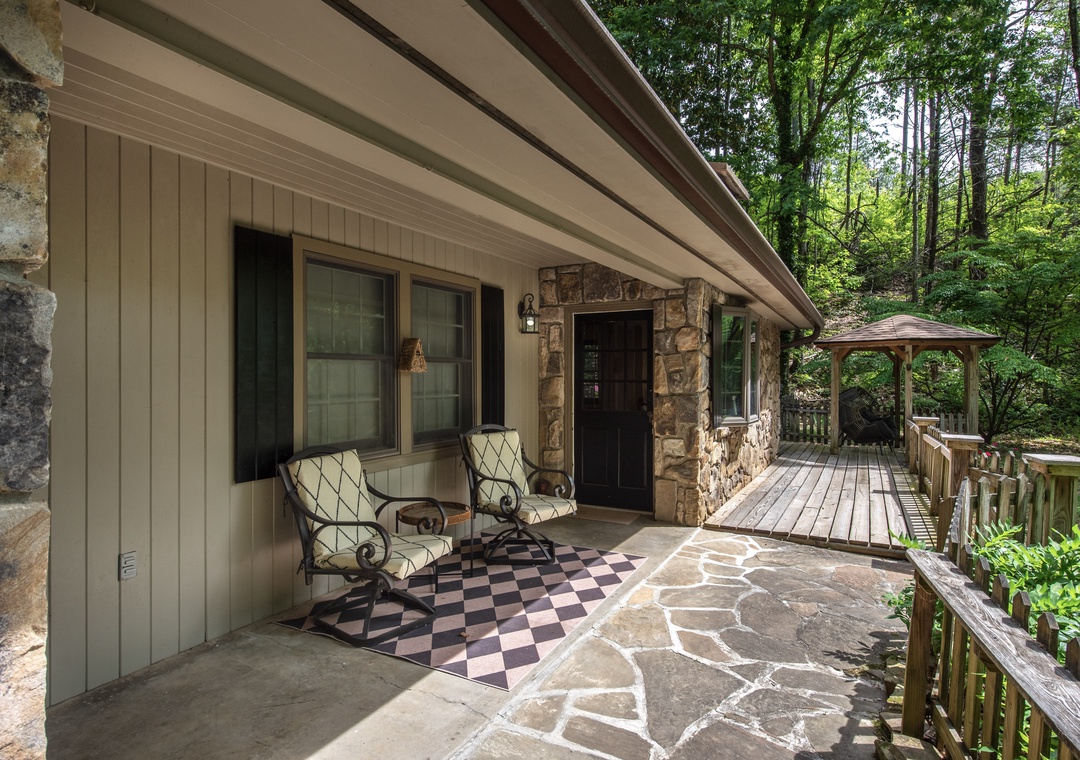 A charming stone-accented entrance welcomes guests, with a spacious covered porch featuring comfortable seating and a peaceful wooded backdrop—your perfect retreat for morning coffee or evening relaxation.