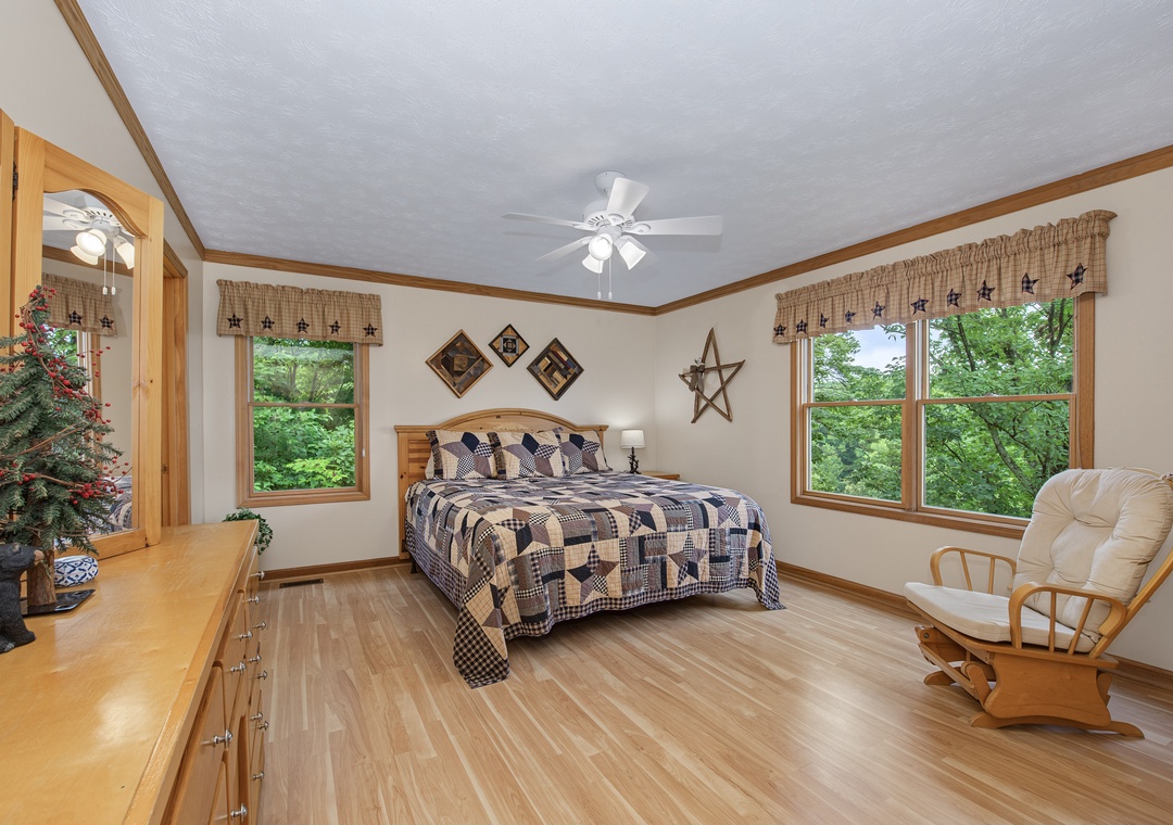 Picture yourself waking in this bright bedroom, wrapped in quilted comfort as lush forest views greet you. Soft morning light filters through, while the cozy rocker invites relaxation.