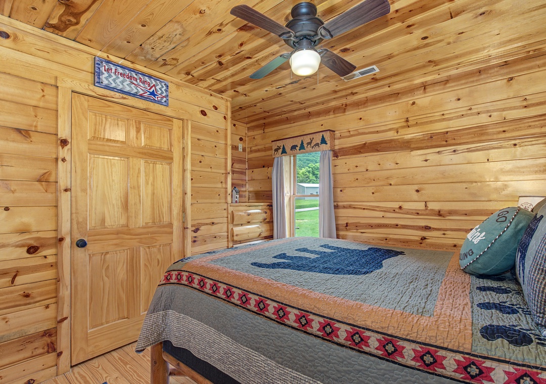 Unwind in this cozy cabin-style bedroom with warm knotty pine walls, a ceiling fan overhead, and a window framing peaceful garden views—your perfect retreat after a day of adventure.