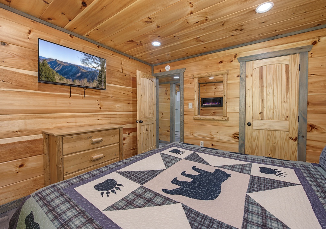 Settle into your cozy cabin bedroom featuring warm pine walls, a wall-mounted TV with mountain views, and a charming bear-themed quilt. Your peaceful retreat includes ample storage and a private entrance.