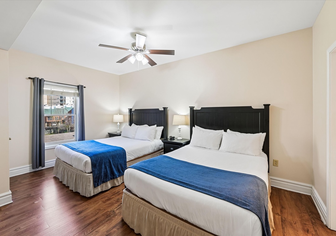 Rest in comfort with two queen beds featuring crisp linens and plush blue throws, while warm wood floors and gentle ceiling fan create your perfect retreat.