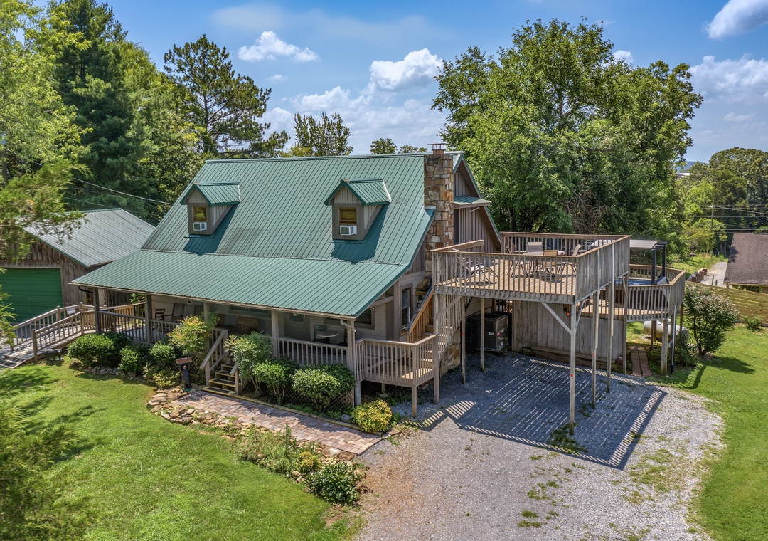 Rustic cabin-style property with green metal roof, wraparound deck, stone chimney, and surrounding mature trees in a peaceful residential setting.