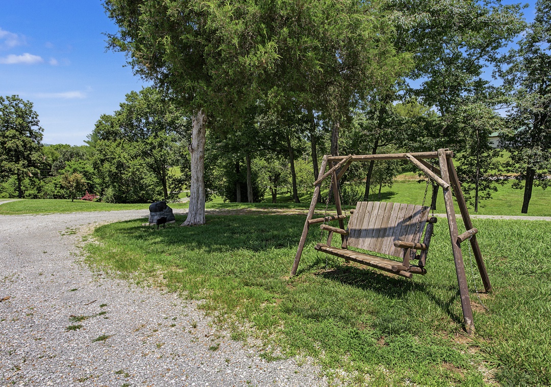 The property features a rustic outdoor swing set on spacious green lawns surrounded by mature shade trees and gravel pathways, offering a peaceful rural setting.
