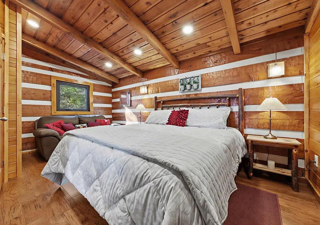 Sink into this plush king-size bed in a charming cabin bedroom with warm wood-paneled walls and vaulted ceilings. The cozy reading chair and rustic touches create your perfect mountain retreat.