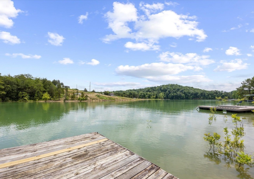 Peaceful lake with wooden dock surrounded by lush hills and clear blue skies in the surrounding area.