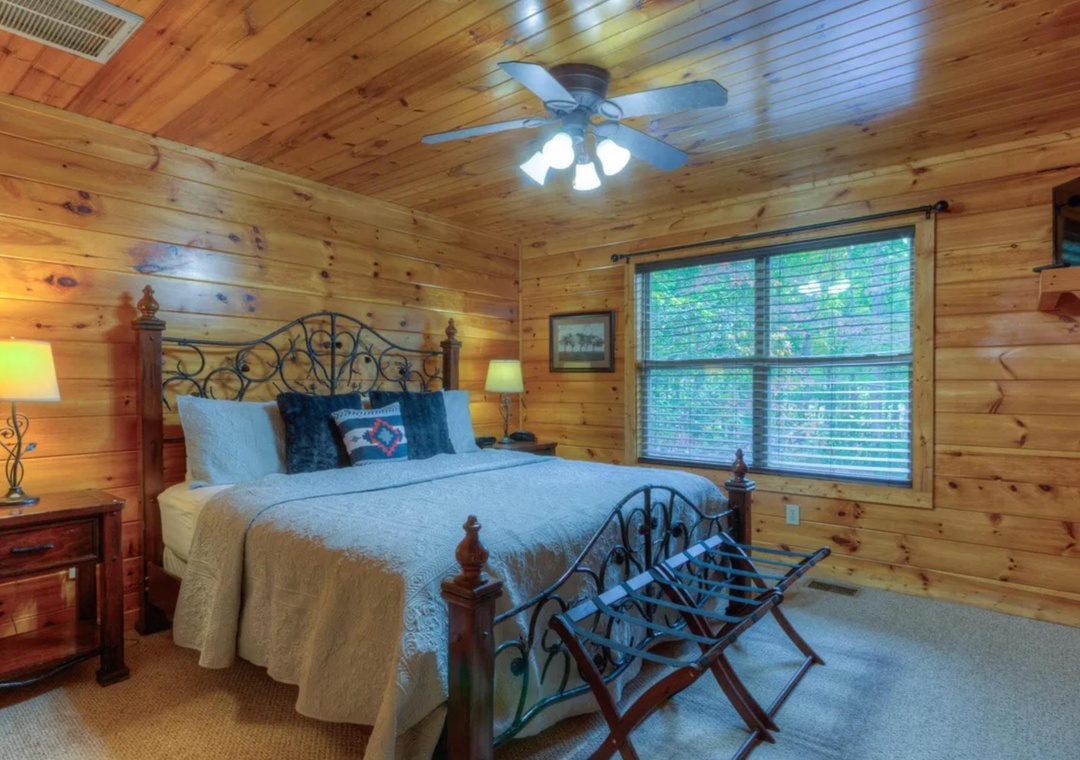 Step into your peaceful retreat with warm pine walls, a plush queen bed with decorative linens, and natural light filtering through blinds. A ceiling fan keeps you cool while the cozy rocking chair invites relaxation.