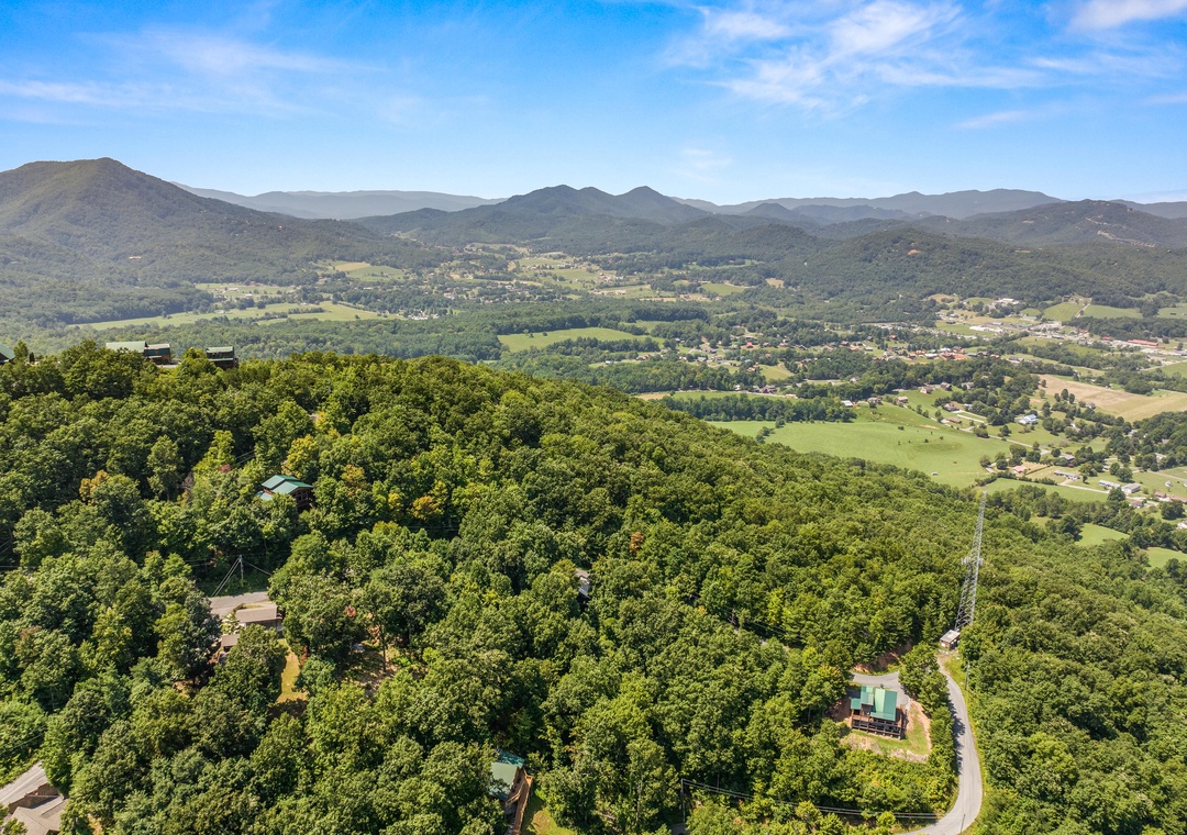 Sweeping aerial view of a mountain valley retreat surrounded by dense forest and rolling peaks. The property sits nestled among trees with scenic vistas of rural countryside and distant mountain ranges.