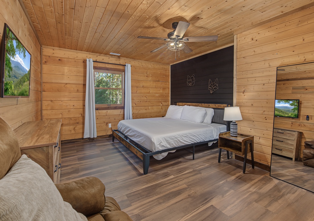 Unwind in your spacious bedroom featuring warm wood paneling, a plush king bed, and cozy seating. Natural light streams in as a ceiling fan keeps you comfortable year-round.