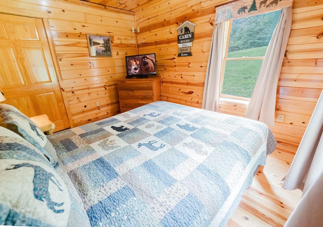 Drift off in this cozy cabin bedroom with warm wood walls, a soft quilted bed, and a window framing peaceful green hills—your private mountain retreat awaits.