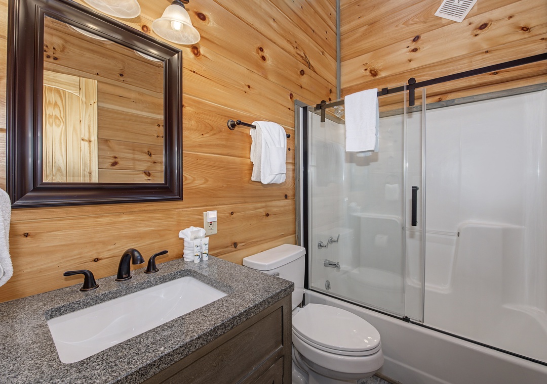 Unwind in your private bathroom featuring a sleek granite vanity, modern fixtures, and a spacious shower-tub combo, all set against warm, natural wood walls for a cozy retreat.