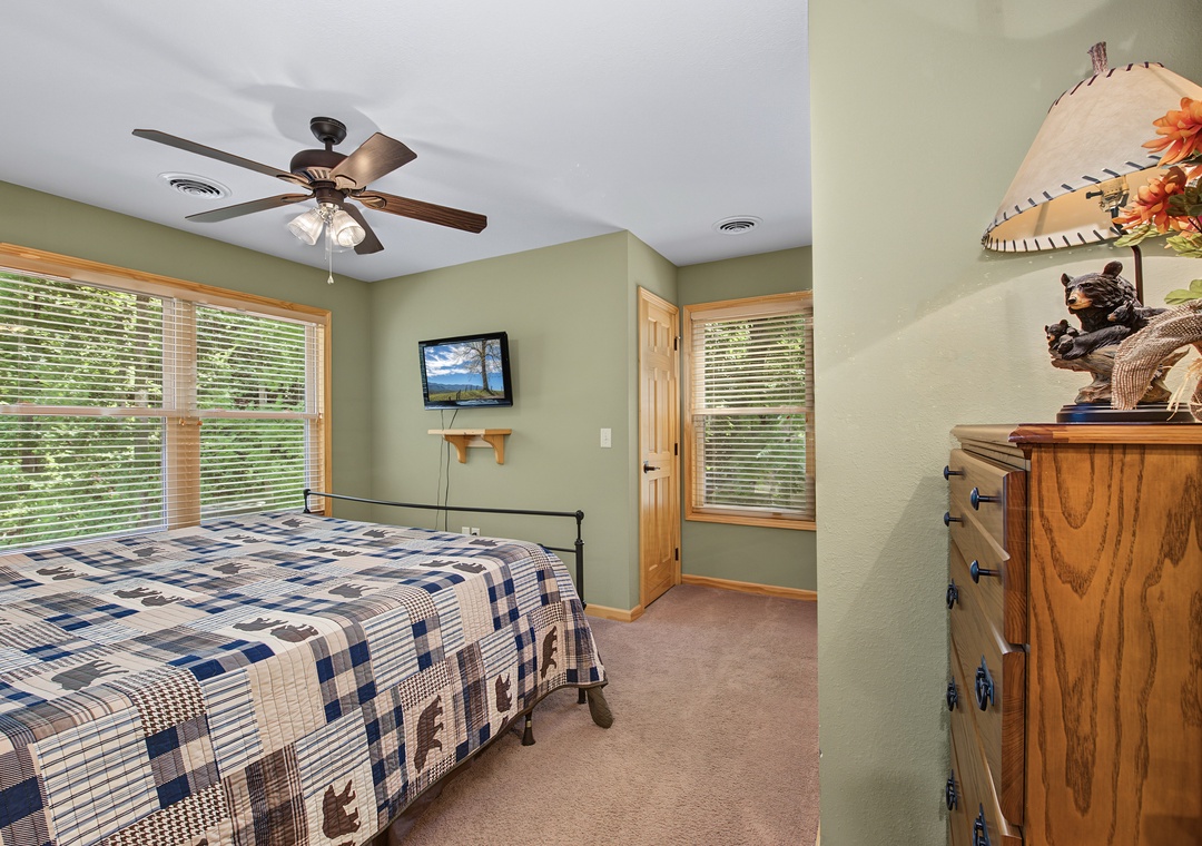 Your serene bedroom features plaid bedding, a ceiling fan for comfort, and bright windows framing peaceful greenery. Warm wood accents and soft sage walls create a cozy retreat.