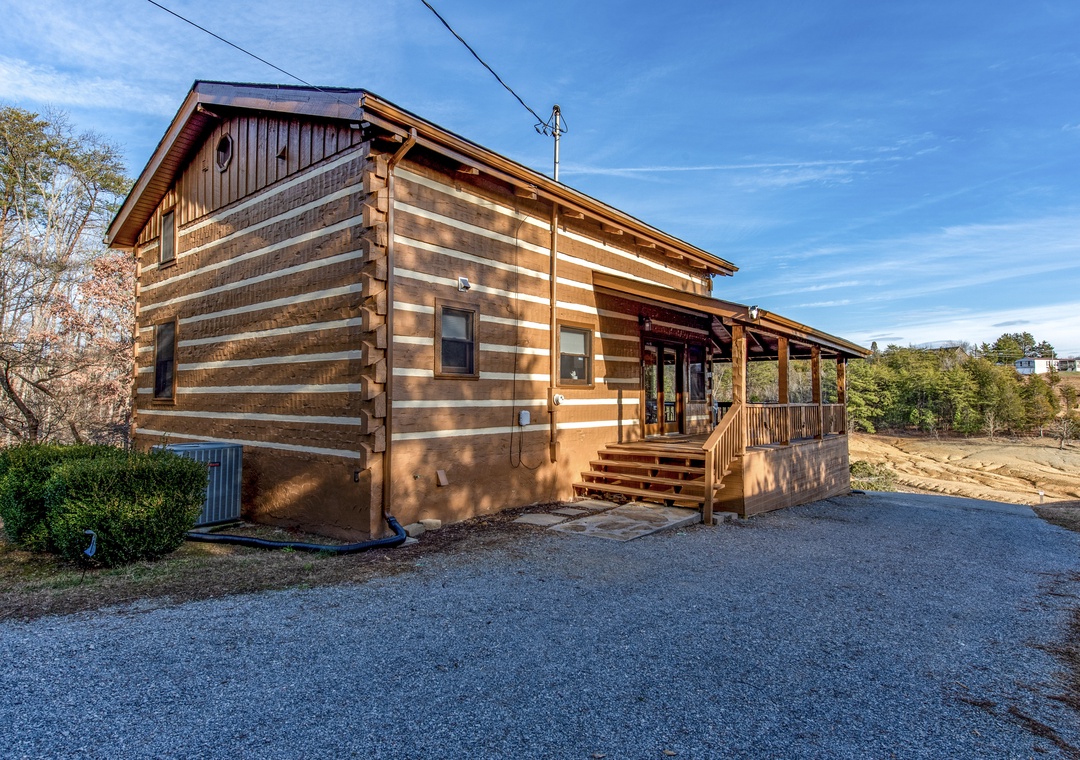 Charming log cabin exterior featuring rustic wood siding, covered porch, and paved parking area surrounded by peaceful countryside.
