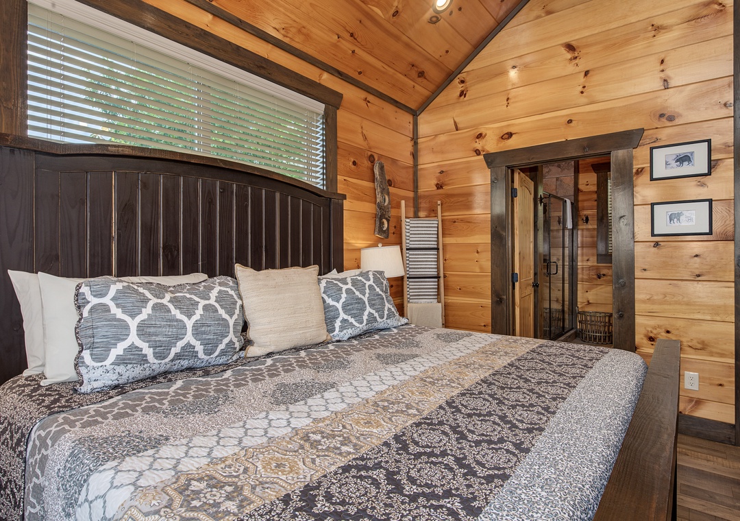Your cozy cabin bedroom features rustic wood walls and vaulted ceilings that create a warm, mountain retreat atmosphere. A plush bed awaits beneath natural light filtering through the window.