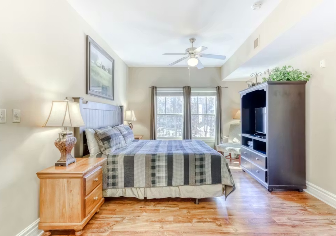 Unwind in your cozy bedroom featuring a plaid-dressed bed, warm wood furnishings, and entertainment center for peaceful evenings.