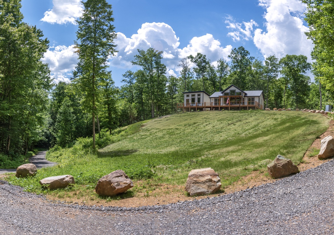 A modern vacation home nestled on a private hillside, surrounded by lush forest and framed by dramatic summer clouds—a peaceful retreat in nature.