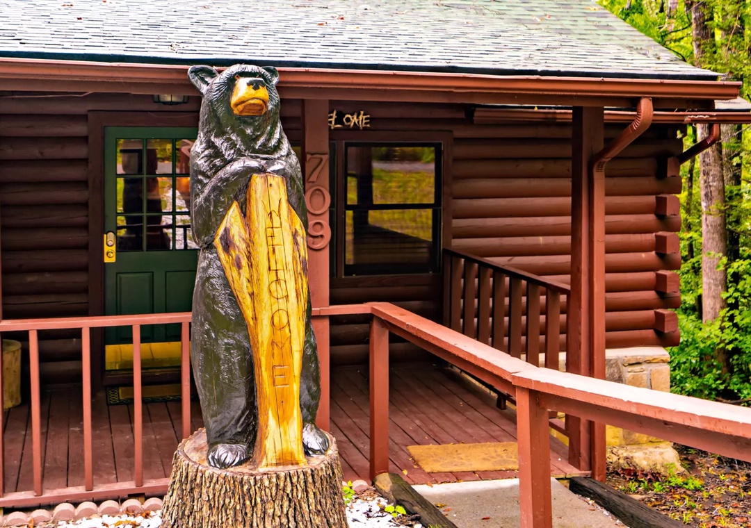 Charming log cabin entrance with rustic bear sculpture and covered deck, surrounded by lush forest setting.