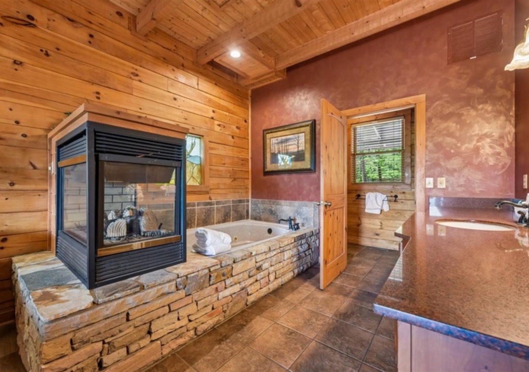 Unwind in your private spa-like bathroom with a soaking tub framed by a cozy two-sided fireplace, warm wood accents, and natural light streaming through the windows.