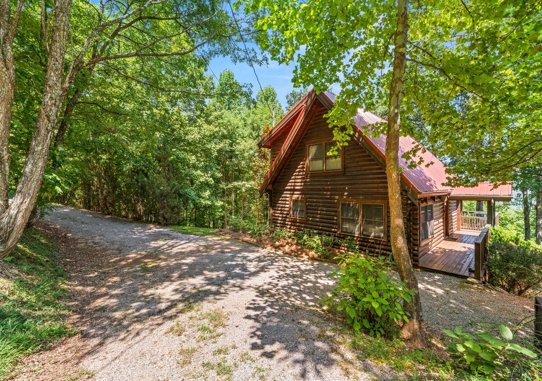 A charming log cabin nestled in a lush forest setting, surrounded by tall trees and accessible via a peaceful gravel drive with convenient parking.