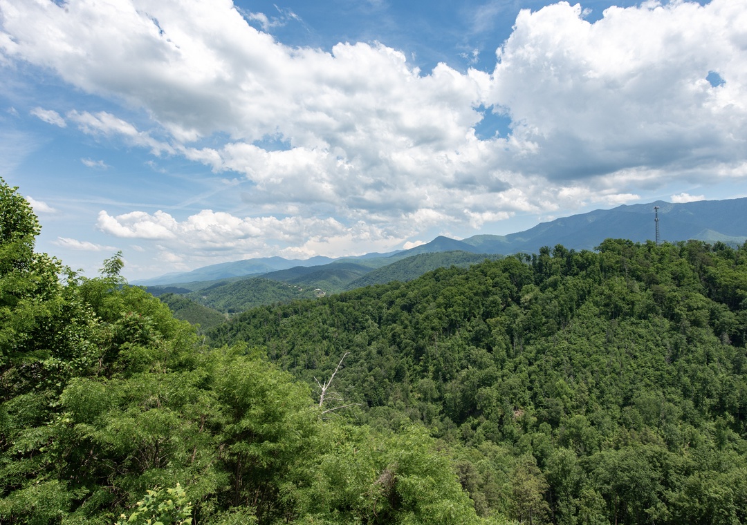 A breathtaking mountain landscape surrounds the property, with rolling forested hills stretching beneath dramatic summer clouds and majestic peaks in the distance.