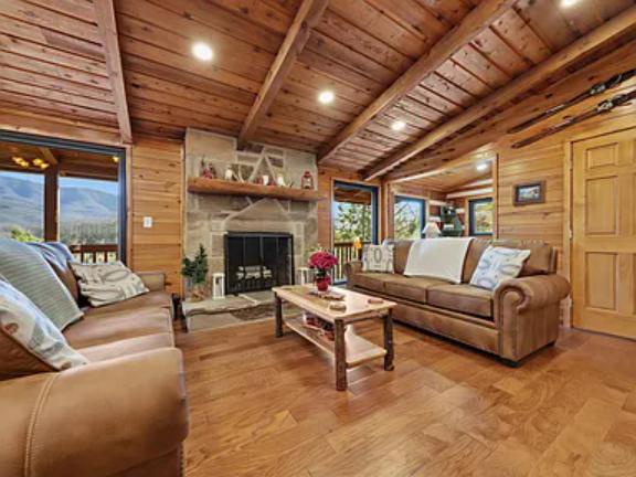 Relax in this warm, wood-beamed living room where exposed timber ceilings, a stone fireplace, and plush seating invite you to unwind while mountain views frame the space through large windows.