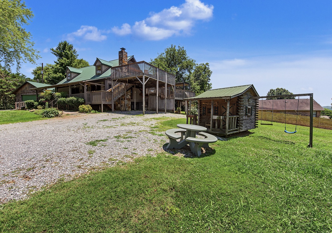 A spacious vacation retreat featuring log cabin architecture with green metal roofing and expansive decks, set on lush grounds with a picnic area, fire pit, and nearby playhouse.