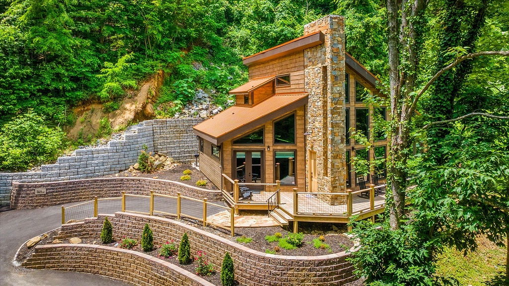 Modern mountain retreat nestled in lush forest with stone chimney, spacious deck, and terraced landscaping—an inviting arrival in a natural woodland setting.
