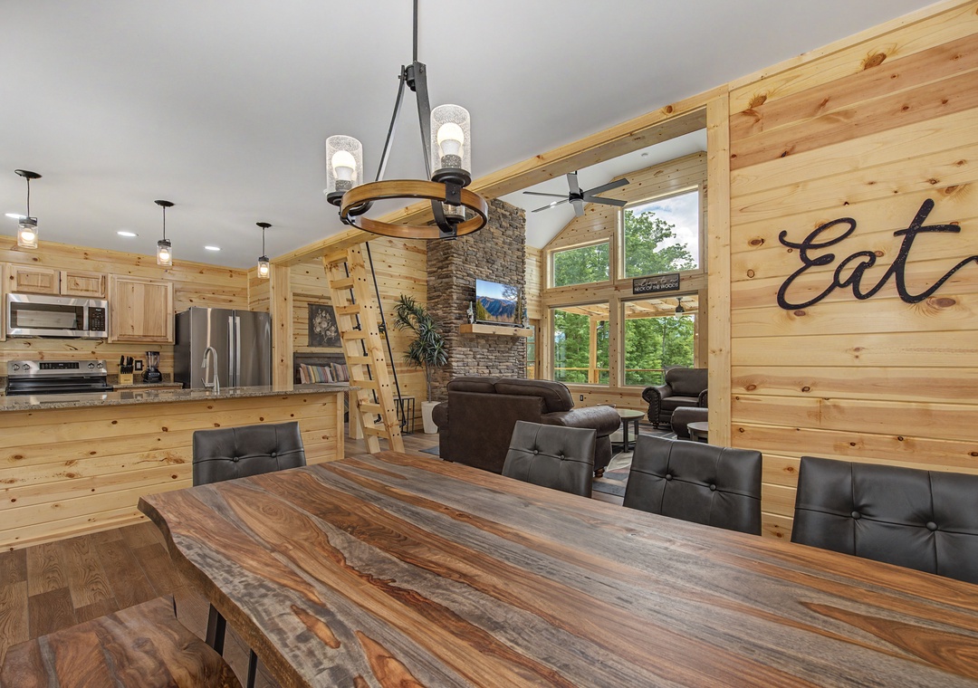 Gather around the handcrafted dining table where rustic charm meets modern comfort. The open-concept design flows from kitchen to living area, with soaring windows framing lush forest views and warm wood details throughout.