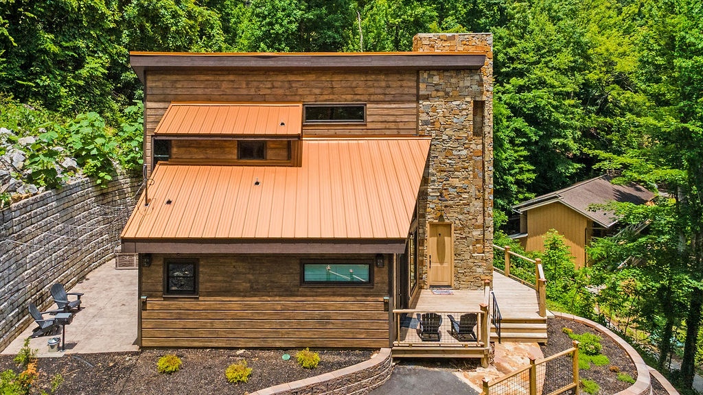 Modern retreat nestled in lush woodland, featuring striking architecture with warm metal accents and natural stone chimney, offering peaceful seclusion and comfort.