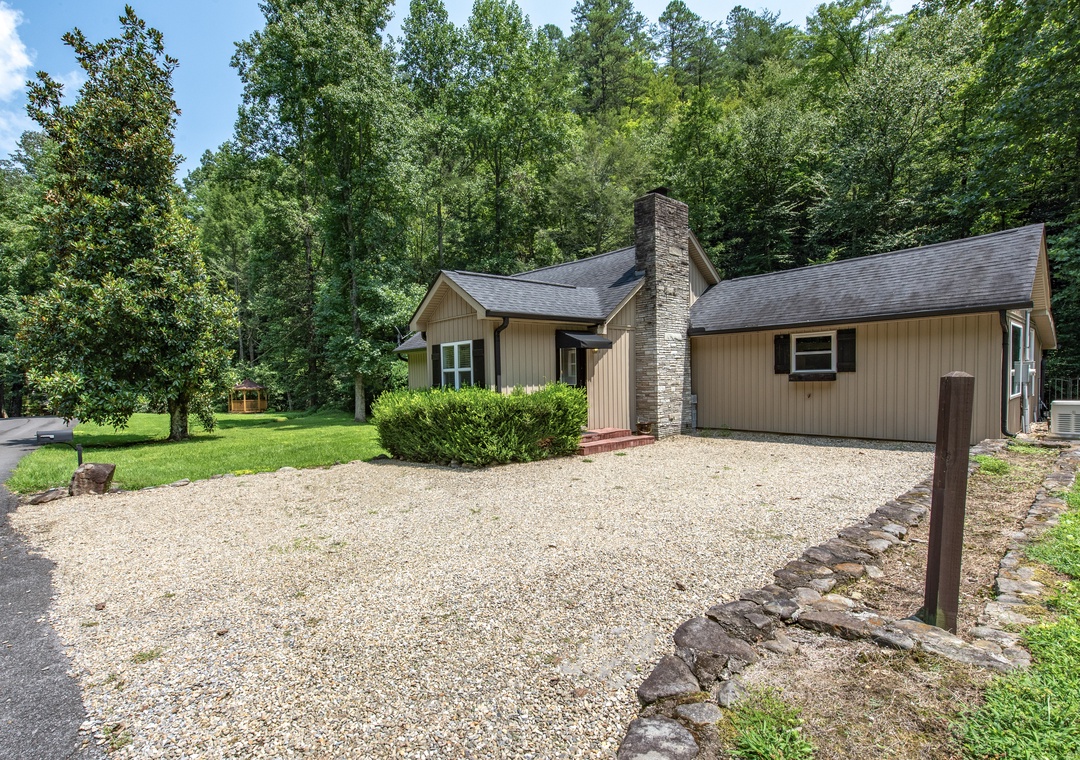 Charming single-story retreat nestled in a lush, wooded setting with a stone chimney and spacious gravel driveway offering plenty of parking.