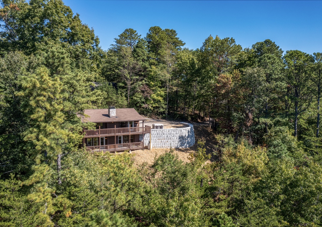 An aerial view captures the property nestled privately among tall pines and hardwoods. The home features spacious wrap-around decks, perfect for enjoying the peaceful, wooded surroundings and clear blue skies.