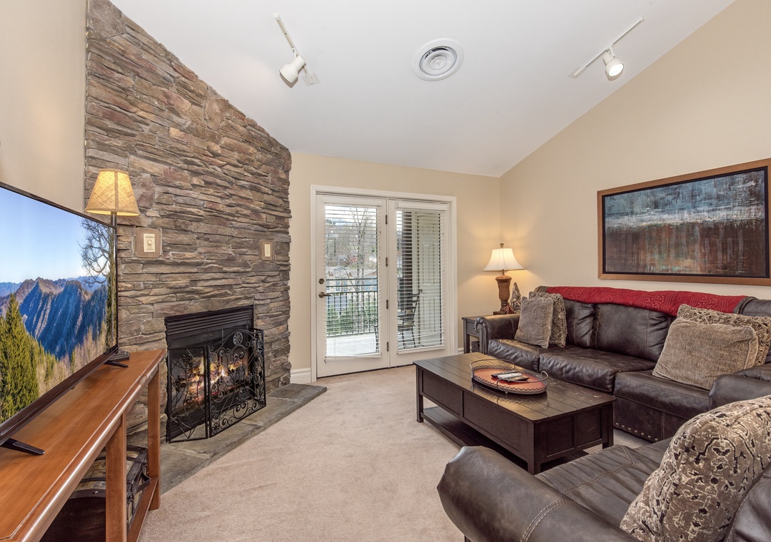 Relax in your cozy living room with stone fireplace, plush leather seating, and mountain views through glass doors to your private balcony.