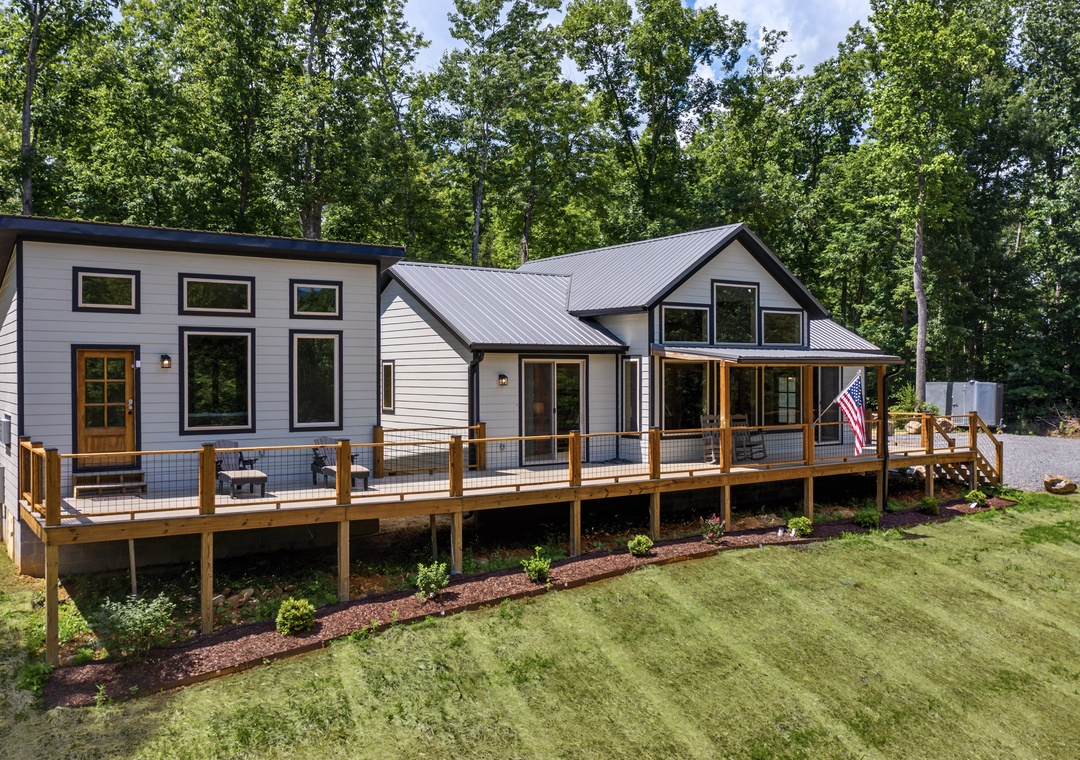 Modern farmhouse exterior featuring metal roofing, expansive windows, and a wraparound deck set among peaceful woods. Well-maintained lawn and landscaping complete the serene forest setting.