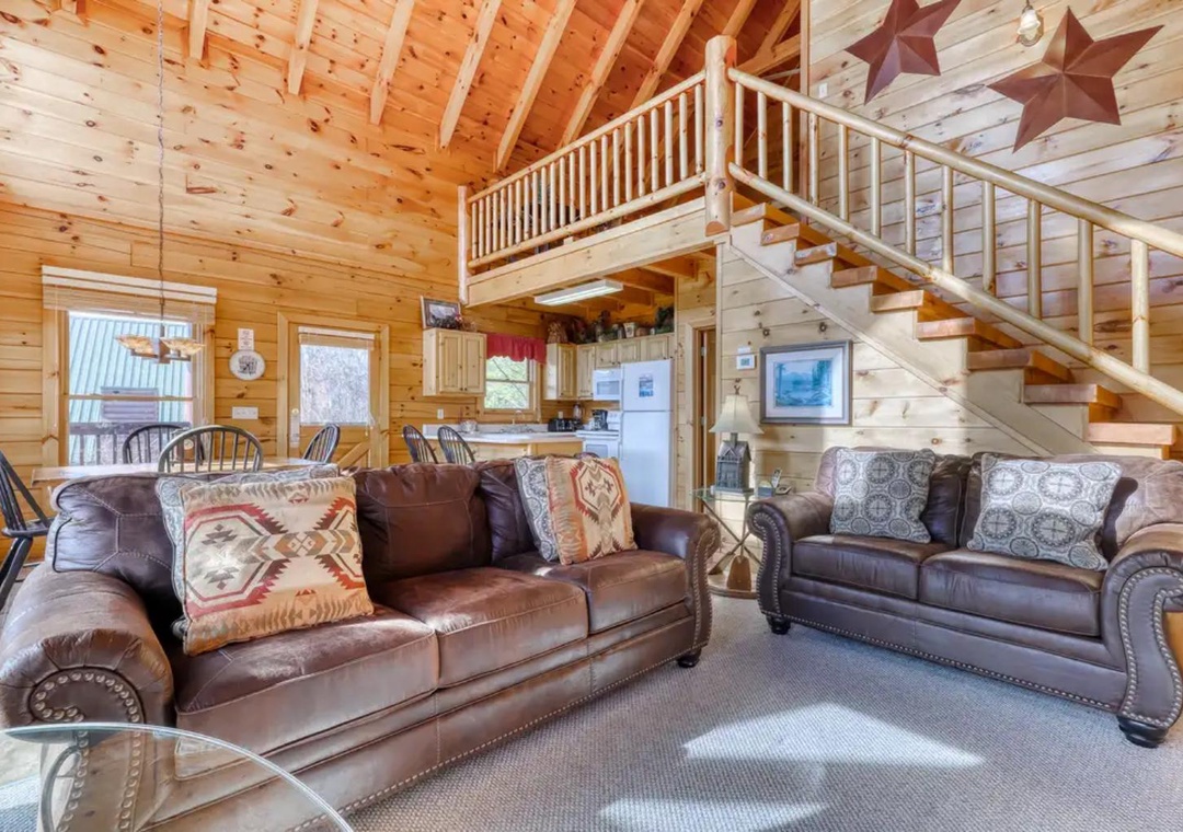 Relax in your spacious cabin living room featuring rich leather couches, warm wood walls, and a soaring cathedral ceiling. The cozy loft above and open kitchen beyond create an inviting mountain retreat.