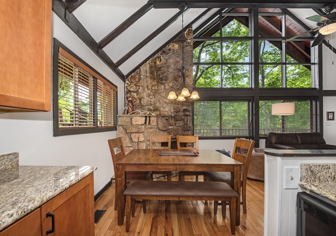 Picture yourself enjoying meals at this rustic dining table beneath soaring vaulted ceilings and exposed beams. Sunlight streams through tall windows, highlighting the charming stone accent wall and warm wood tones throughout your cozy retreat.