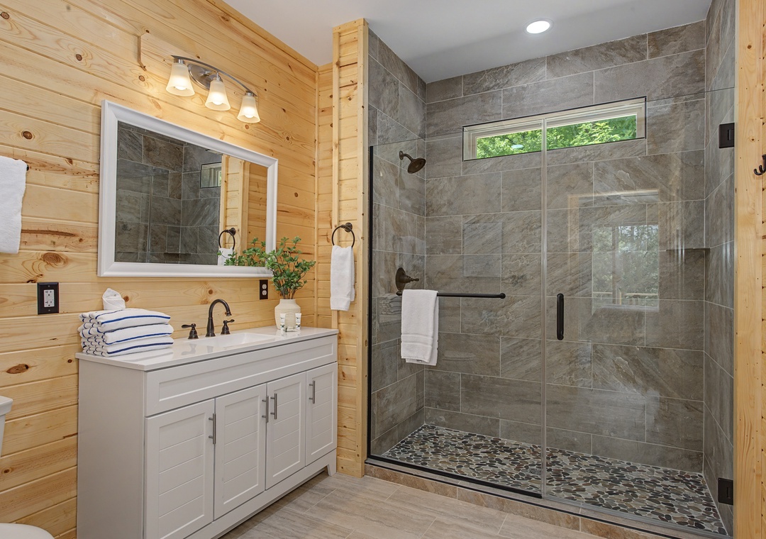 Refresh in your modern bathroom featuring a spacious walk-in shower with natural light and stylish stone tile throughout. Fresh towels and cabin-inspired wood walls create a warm retreat.