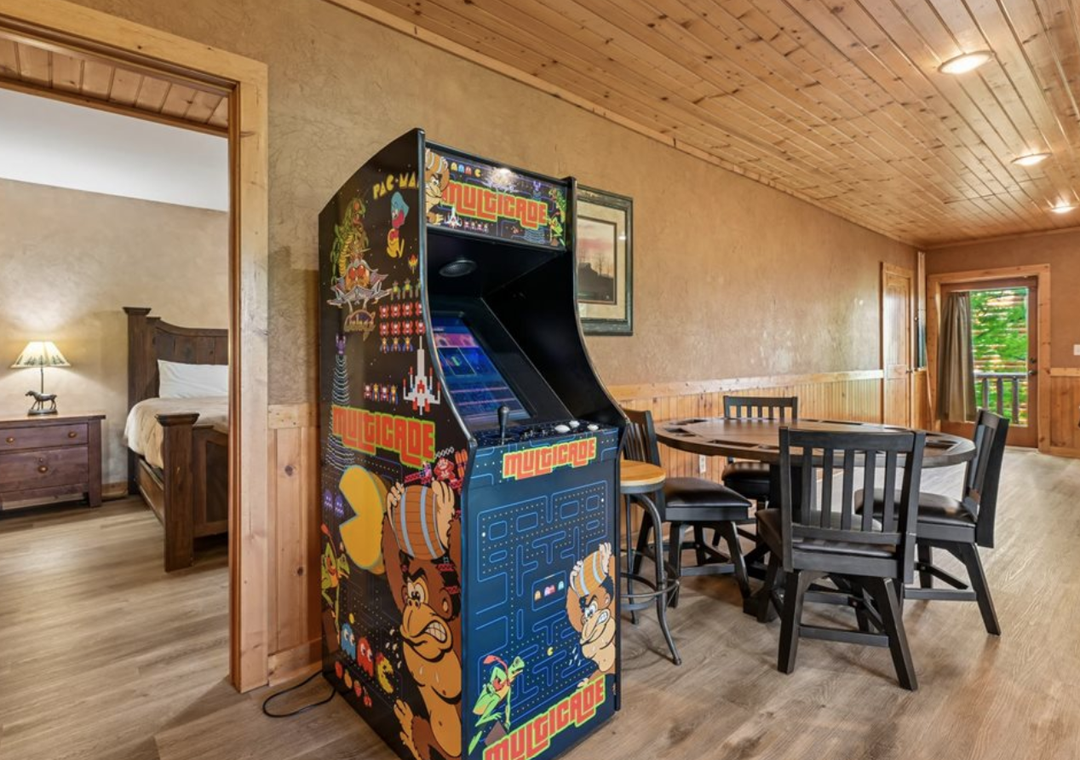 A classic arcade game stands ready for friendly competition, with a cozy dining area just beyond and your bedroom visible through the open doorway—all under warm wood ceilings.