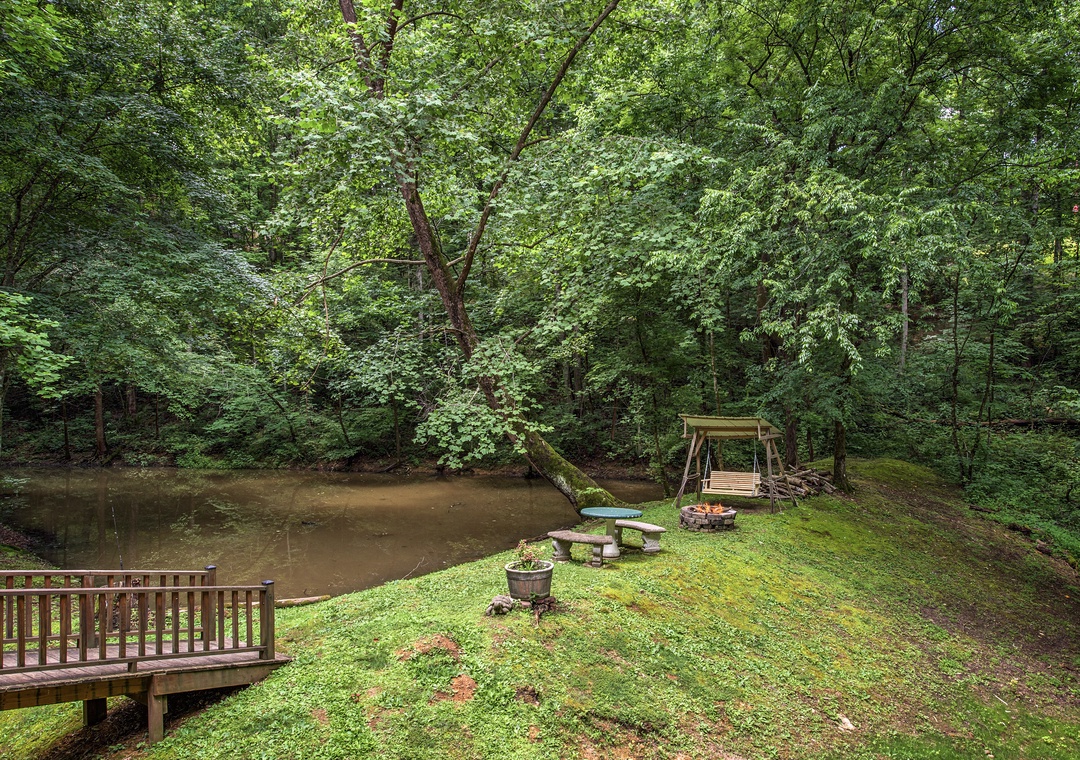 Private waterfront retreat surrounded by lush forest, featuring a tranquil pond, fire pit, picnic area, and scenic deck for peaceful nature escapes.