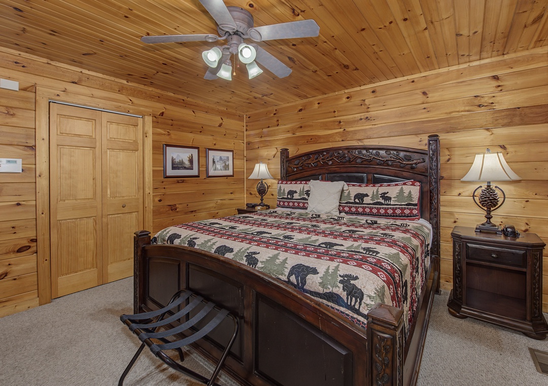 Sink into rustic charm beneath warm wood paneling and a gentle ceiling fan. Your cozy retreat features a beautifully carved bed with wilderness-themed linens, inviting lamps, and convenient bedside storage.