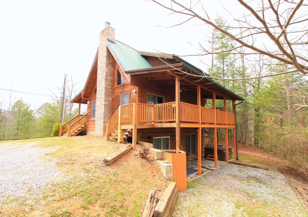Charming log cabin with wraparound deck nestled among trees, featuring stone chimney and green metal roof in peaceful wooded setting.