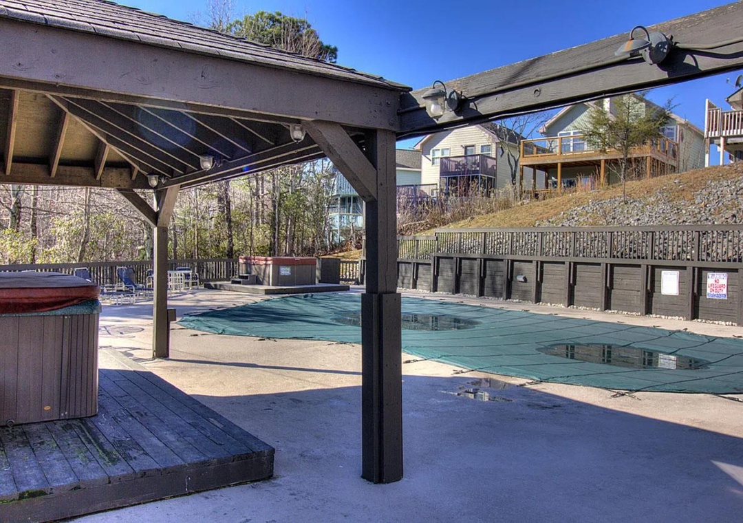 Resort-style pool area with covered seating and hot tub, surrounded by hillside vacation homes under clear blue skies.