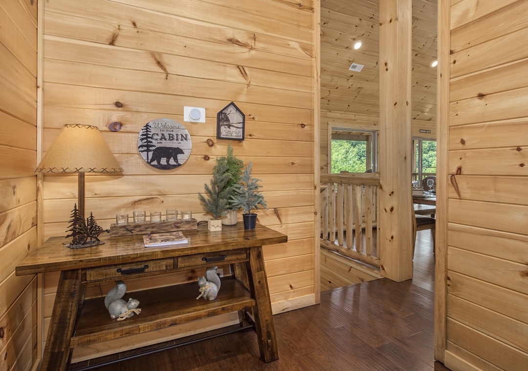 Step inside your cozy cabin retreat where warm wood planks embrace every surface. The rustic console welcomes you with charming forest-inspired décor, while natural light streams through windows framing lush greenery beyond.