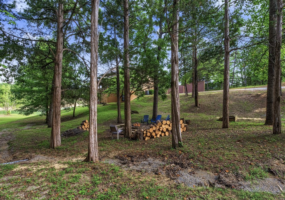 This peaceful wooded area offers a shaded outdoor retreat with seating around a fire pit and stacked firewood, perfect for cozy evenings under the trees on the property grounds.