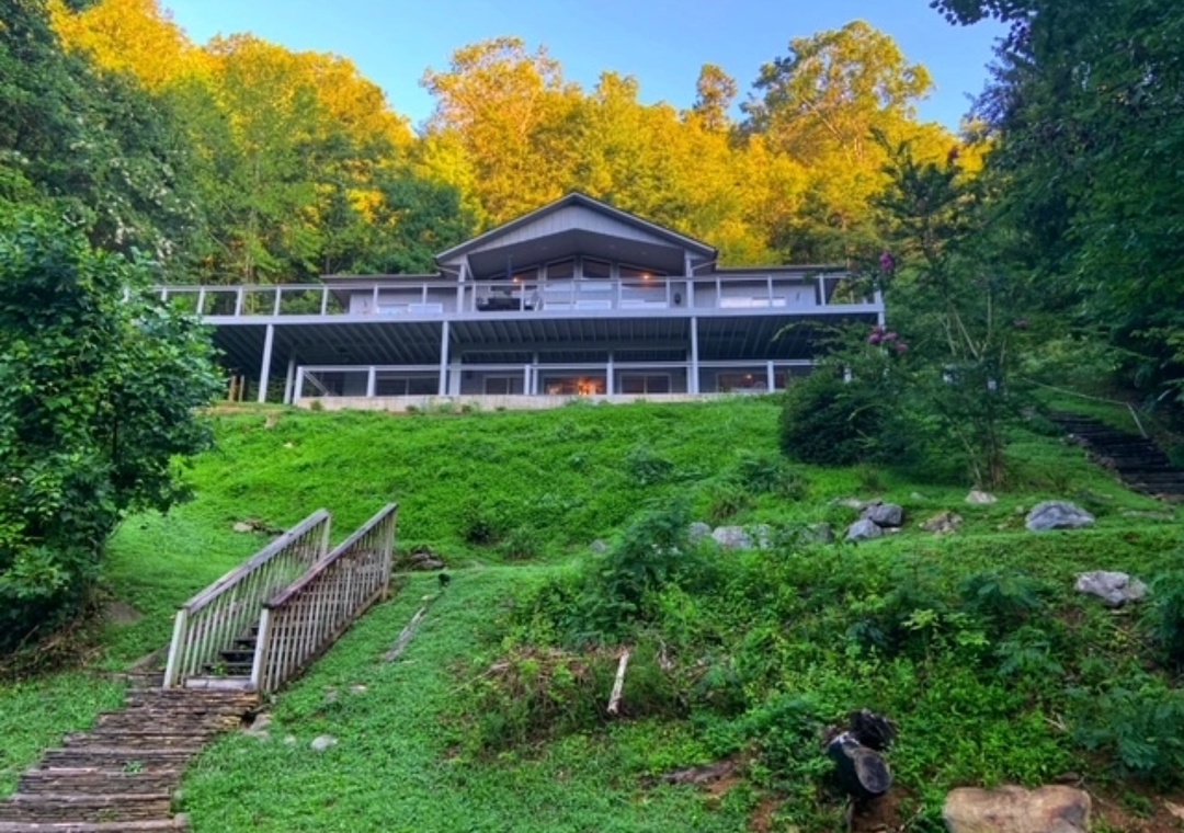 Modern lakeside retreat nestled among vibrant autumn foliage, featuring multi-level decking and natural stone landscaping in a serene wooded setting.