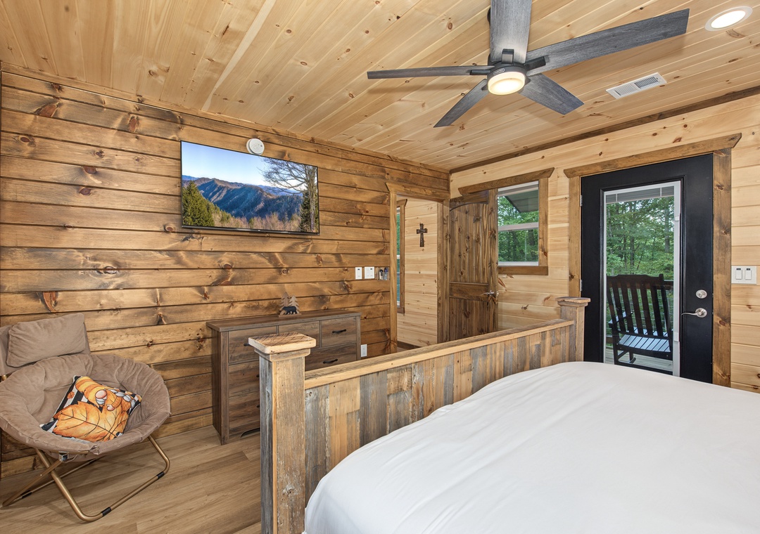 Cozy wood-paneled bedroom with vaulted ceiling, modern fan, and glass door opening to your private balcony surrounded by trees.