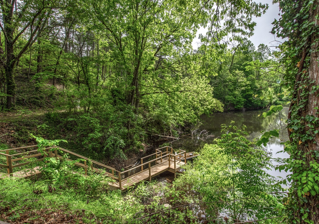 A tranquil wooden footbridge leads to the calm waters edge, surrounded by lush trees and natural beauty that define the peaceful setting of this vacation rental.