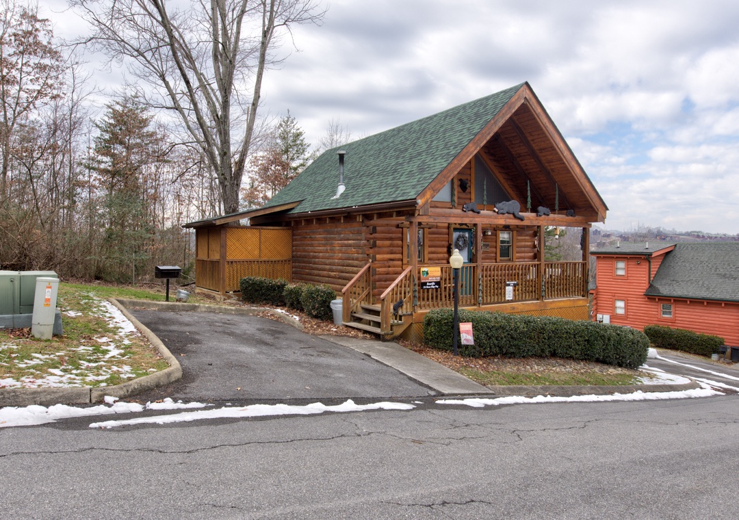 Charming log cabin with wraparound deck nestled among trees, featuring convenient parking and peaceful woodland surroundings.