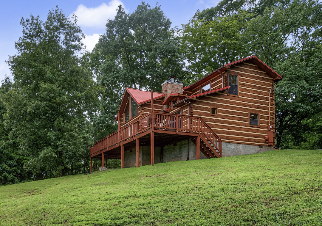 A charming log cabin nestled in a lush wooded hillside, framed by towering trees and open lawn. The property features warm wood siding, a red metal roof, and an inviting multi-level deck perfect for outdoor relaxation.