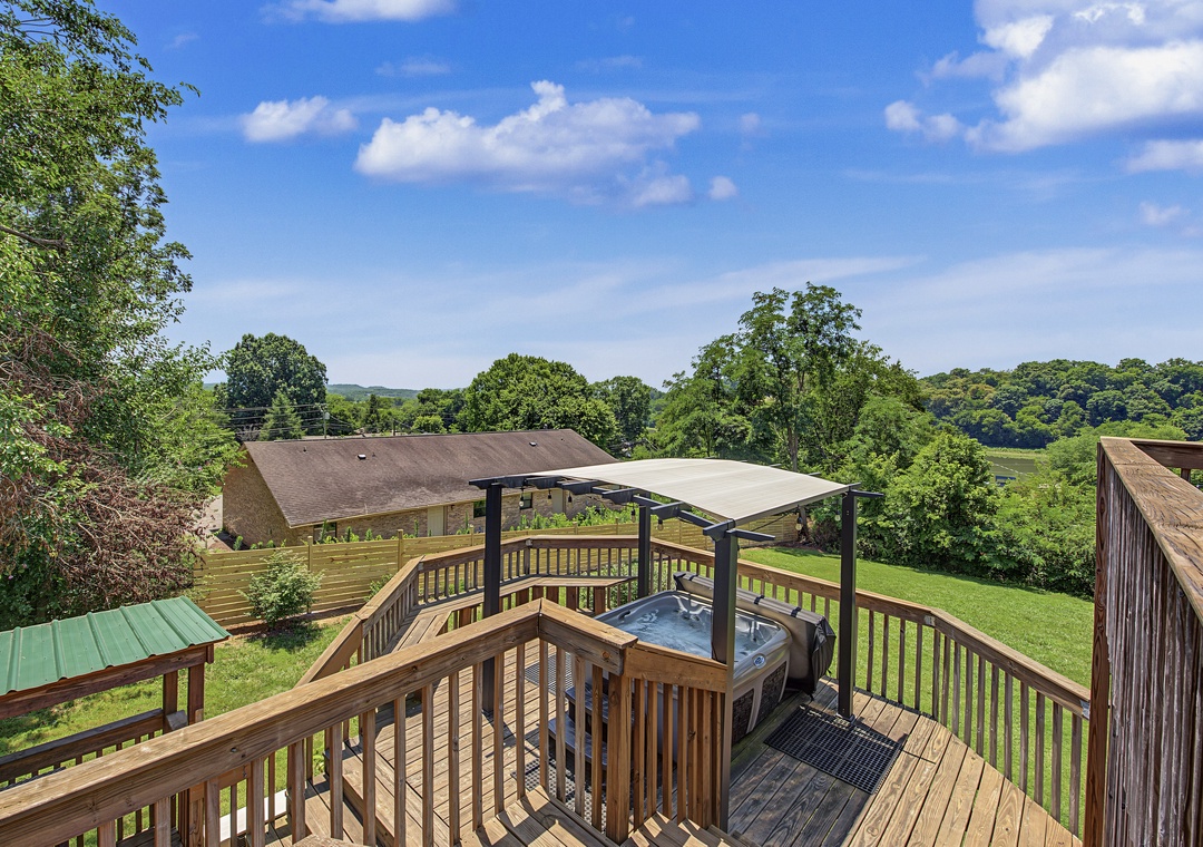 An elevated deck features a private hot tub overlooking peaceful countryside, framed by mature trees and rolling green hills—a perfect spot for relaxing under open skies.